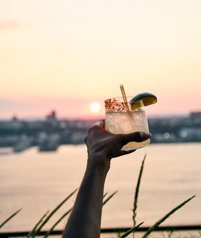Hand holding a cocktail with lemon slice, city skyline and sunset in background.