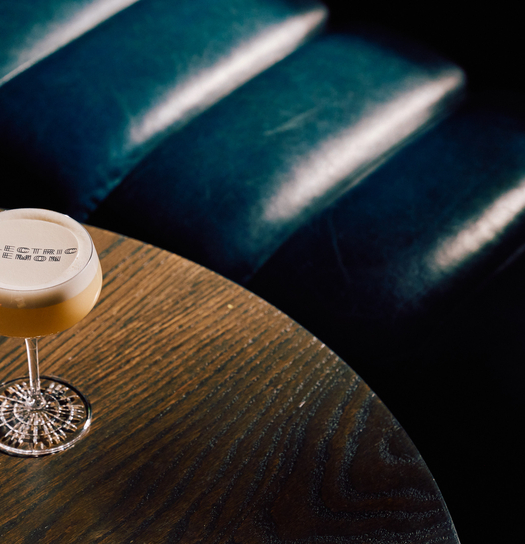 A glass with "Electric Lemon" logo sits on a round wooden table, illuminated by sunlight, with a dark blue leather chair in the background.