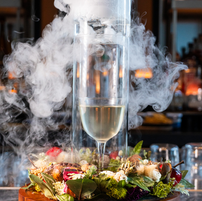 Sparkling wine in a tall glass with smoke, decorative floral arrangement on table, dimly lit bar background.
