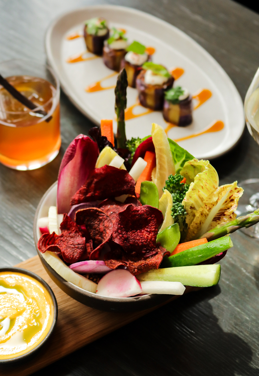 Chic restaurant dish featuring vibrant salad with beet chips, mixed greens, and asparagus, served with a drink and a small appetizer on a dark table.