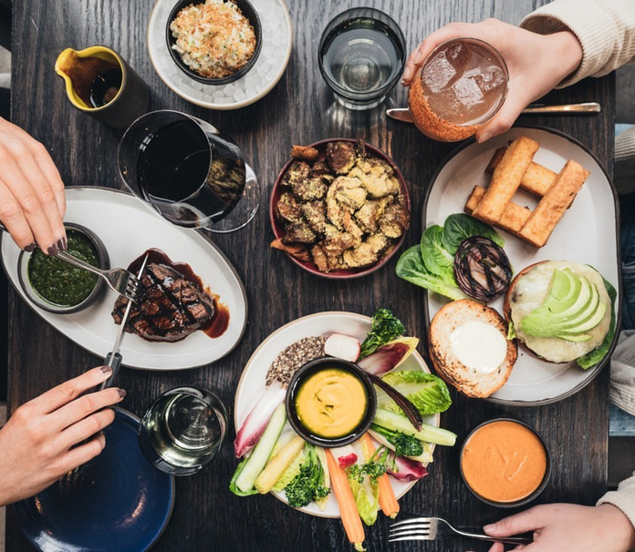A top-down view of a dining table featuring a variety of dishes, including grilled meat, broccoli, carrots, sandwiches, and desserts, with drinks in glasses.