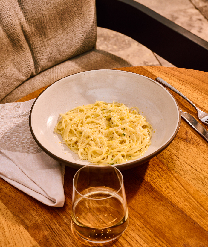 A bowl of pasta with creamy sauce on a wooden table, served with white wine and utensils on a striped napkin.