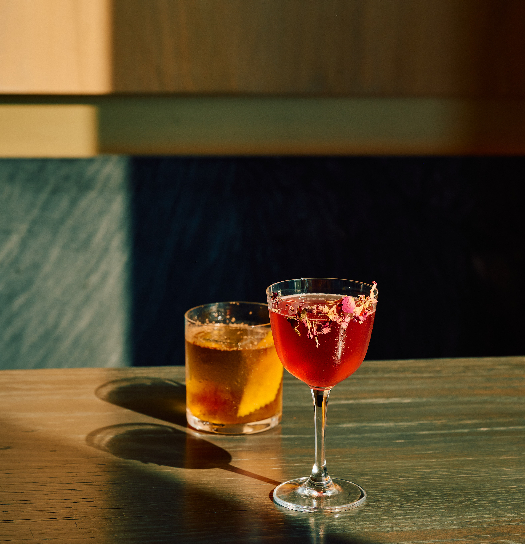 Two cocktails sit on a sunlit wooden table: a red drink in a stemmed glass with a sugared rim in the foreground, and an amber drink in a short tumbler behind it, set against a dark wall.