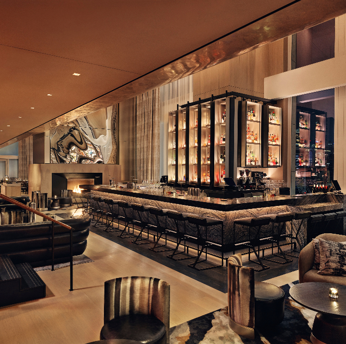A stylish modern bar lounge with a long marble counter, tall backlit liquor shelves, upholstered bar stools, and warm ambient lighting in a high-ceilinged space.