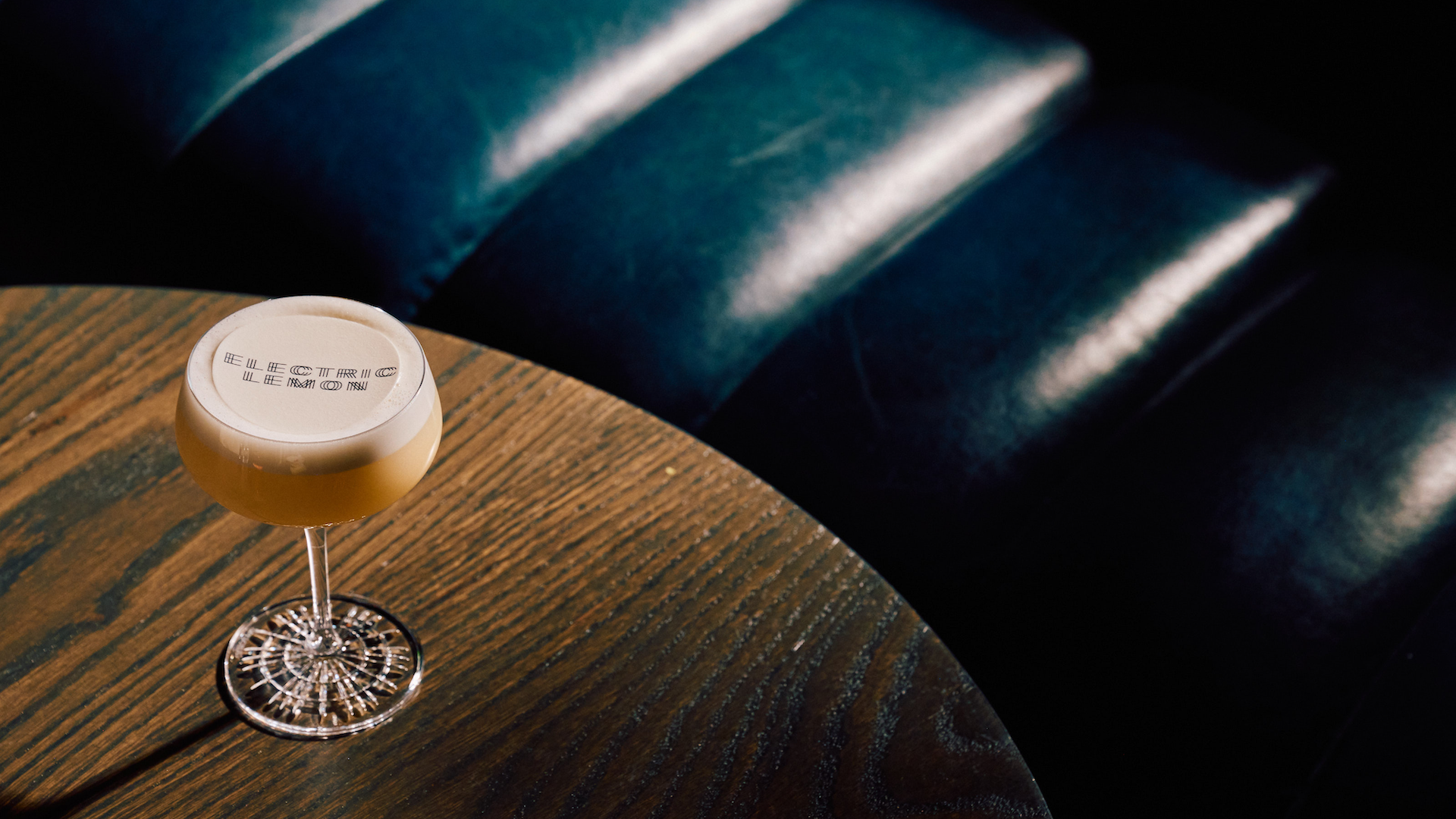 A frothy cocktail in a coupe glass, its foam topped with an “Electric Lemon” logo print, sits on a round dark wood table beside a deep teal leather banquette catching streaks of light.