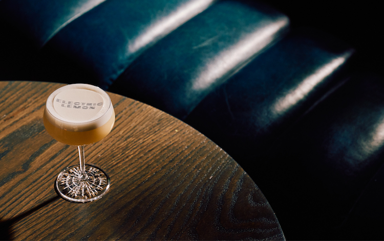 A frothy cocktail in a coupe glass, its foam topped with an “Electric Lemon” logo print, sits on a round dark wood table beside a deep teal leather banquette catching streaks of light.